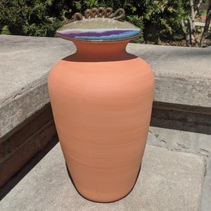 May include: A terracotta-colored ceramic vase with a decorative lid. The lid features a swirl design with shades of green, purple, and blue. The vase has a wide base and a narrower neck, suitable for home decor.