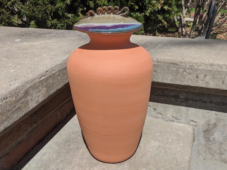 Puede incluir: Un jarr&oacute;n de cer&aacute;mica color terracota con tapa decorativa. La tapa presenta una banda de colores morado, azul y verde, y un asa de metal retorcido. El jarr&oacute;n es cil&iacute;ndrico con una abertura ligeramente ensanchada, ideal para decoraci&oacute;n.