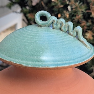 Puede incluir: Olla de cer&aacute;mica hecha a mano con tapa turquesa y asa decorativa en espiral. La olla tiene una base de color terracota y una tapa turquesa con superficie texturizada. El asa en espiral a&ntilde;ade un toque &uacute;nico al dise&ntilde;o.