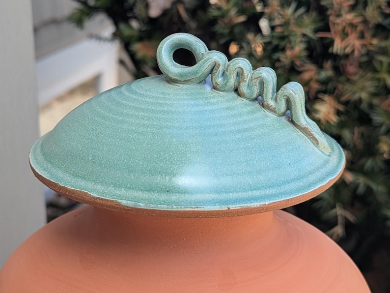 Puede incluir: Olla de cer&aacute;mica hecha a mano con tapa turquesa y asa decorativa en espiral. La olla tiene una base de color terracota y una tapa turquesa con superficie texturizada. El asa en espiral a&ntilde;ade un toque &uacute;nico al dise&ntilde;o.