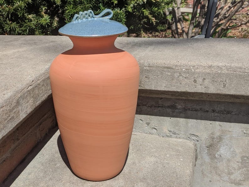 Puede incluir: Un jarr&oacute;n de cer&aacute;mica grande, color terracota, con tapa azul claro. La tapa tiene un asa decorativa. El jarr&oacute;n es cil&iacute;ndrico con la parte superior ligeramente ensanchada. El jarr&oacute;n est&aacute; sobre una superficie de hormig&oacute;n.