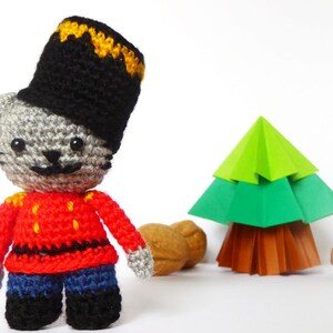 Op de afbeelding: Een grijze gehaakte kat met een zwarte en gele gehaakte hoed en een rood gehaakt jasje met gele accenten. De kat staat voor een groene origami kerstboom en bruine walnoten.