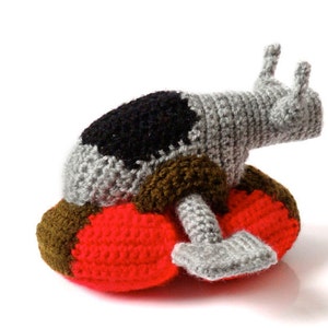 May include: A handmade crochet slug toy in gray, black, brown, and red. The slug has a gray body with a black patch, brown and red base, and two gray antennae. The toy is made of yarn and is on a white background.