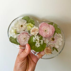 May include: Clear resin coasters with embedded dried flowers. The coasters feature a variety of pink, white, and yellow flowers, along with green leaves. The coasters are round and appear to be handmade.