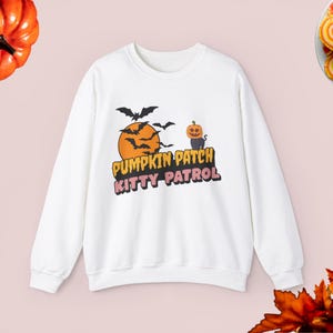 Puede incluir: Sudadera blanca con un estampado gráfico de un gato negro con cabeza de calabaza, murciélagos volando alrededor de una luna llena y el texto "Pumpkin Patch Kitty Patrol".
