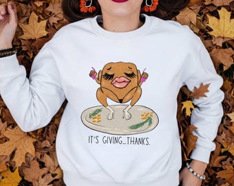 Può includere: Felpa bianca con una grafica umoristica di un tacchino su un piatto. Il tacchino ha lunghe ciglia, rossetto rosa e tiene lo smalto rosa. Il testo "IT'S GIVING...THANKS." è sotto. La modella indossa jeans blu.