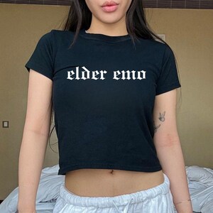 May include: Black crop top with the text "elder emo" in white gothic font. The short-sleeved shirt is made of a soft material.