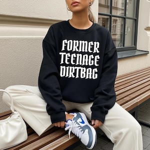 Könnte beinhalten: Schwarzes Sweatshirt mit dem weißen Text "FORMER TEENAGE DIRT BAG" in fetter, gotischer Schrift. Das Sweatshirt wird mit cremefarbener Hose und weißen Turnschuhen mit blauen Akzenten getragen. Eine weiße Handtasche ist ebenfalls zu sehen.