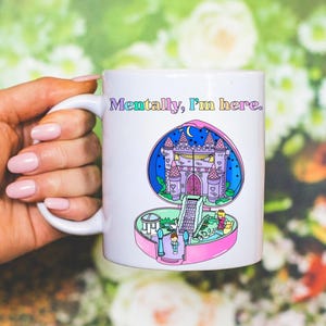 Op de afbeelding: Witte keramische mok met een kleurrijke illustratie van een kasteel in een hartvormige compact. De mok heeft de tekst "Mentally, I'm here." in regenboogkleuren. De mok wordt vastgehouden door een hand met roze nagellak.