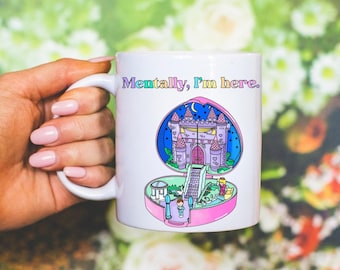 May include: White ceramic mug with a colourful illustration of a castle inside a heart-shaped compact. The mug has the text "Mentally, I'm here." printed in rainbow colours. The mug is held by a hand with pink nail polish.