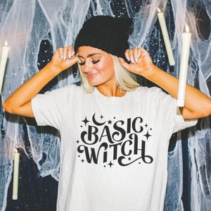May include: Off-white t-shirt with the text "BASIC WITCH" in black script, adorned with stars and a crescent moon. The person in the image is wearing a black beanie. The theme is Halloween.