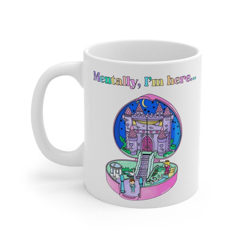Op de afbeelding: Witte keramische mok met een kleurrijke illustratie van een miniatuurkasteel in een roze ovaal. De mok heeft de tekst "Mentally, I'm here..." in een pastelkleurige lettertype. De handgreep zit aan de linkerkant.