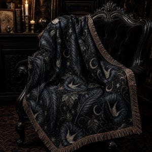 Può includere: Una coperta tessuta di colore scuro con un motivo ripetuto di grandi piume, fiori di gloria mattutina, lune crescenti e piccole stelle. La coperta ha una superficie strutturata ed è rifinita con un bordo decorativo a frange.