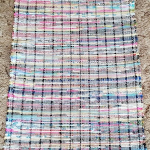 May include: A rectangular woven rug with a plaid pattern. The rug features a mix of colors including pink, blue, white, and black. The rug is made of woven fabric and is suitable for use as a floor covering.