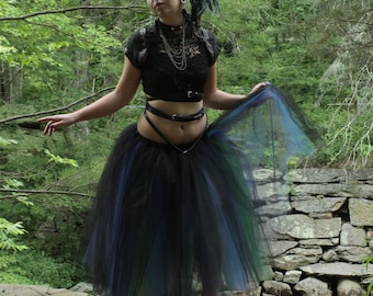 May include: A woman with blue hair and a black crop top is wearing a black tulle skirt with green and blue accents. She is standing in a forest setting with a stone wall behind her.