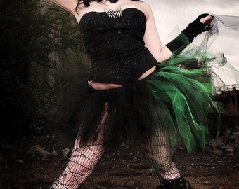 May include: A woman wearing a black corset top, a black and green tulle skirt, black fishnet stockings, and black boots. She has blue hair and is posing in front of a gray and brown background.