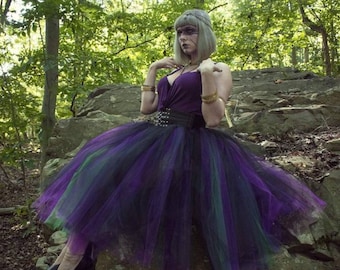 May include: A woman wearing a purple and green tulle skirt with a black belt and a purple top. She is standing in a forest setting.