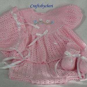 May include: A pink crocheted baby outfit set with a dress, hat, and booties. The dress has a white lace trim and a floral embroidery design. The set is tied with white satin ribbons.