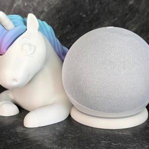 Peut inclure: Une figurine de licorne en céramique blanche avec une crinière et une corne bleues et violettes, à côté d'un haut-parleur intelligent blanc avec un revêtement en tissu maillé gris.