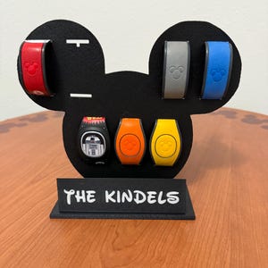 Op de afbeelding: Een zwarte Mickey Mouse-vormige displaystandaard met verschillende kleurrijke MagicBands. De bandjes zijn rood, grijs en blauw, met een Star Wars-thema band en twee oranje en gele bandjes. De standaard heeft een basis met de tekst "THE KINDELS".
