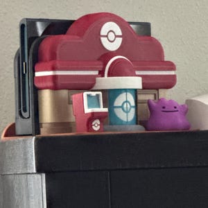 May include: A red and white Pokemon Center playset with a blue and white cylindrical structure and a purple Ditto figure. The playset features a red building with a white Pokeball symbol.