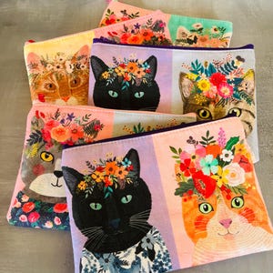 May include: A selection of zipped pouches with cat portraits wearing floral crowns. The cats are depicted in orange, black, and tabby fur, set against pastel backgrounds. These pouches are suitable for carrying small items. Each pouch has a zip closure.