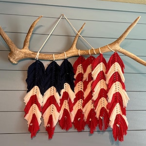 May include: A decorative wall hanging featuring a deer antler base with a macrame design. The macrame is crafted in red, white, and navy blue, resembling a flag. The piece is mounted on a light blue, horizontal-slat wall.