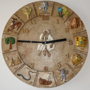 May include: A beige circular clock face with Roman numerals and various illustrations around the edge. The illustrations include a deer, a tree, a tent, a snake, a boat, a donkey, a building, a flower, a bird, and a paint palette. The clock face has a black hand pointing to the number 12.
