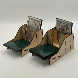 May include: Two wooden card holders with a natural finish. Each holder has a green base and a slot for holding cards. The holders are designed to display and organise trading cards, with cards visible in the slots.