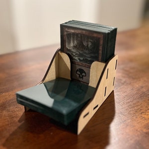 May include: A wooden card holder with a skull design. The holder has a slot for a deck of cards and a compartment for a card box. The holder is made of light brown wood.
