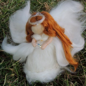 Może przedstawiać: Ręcznie robiona figurka anioła z białą sukienką i dużymi białymi skrzydłami. Anioł ma długie, rude włosy i trzyma błyszczący kryształ. Halo i mały kryształowy kolczyk zdobią głowę anioła. Figurka stoi na zielonej trawie.