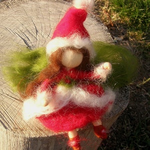 May include: A handmade felt doll wearing a red Santa hat with white trim, a red dress with white trim, and green wings. The doll has a white face and brown hair.