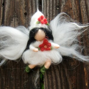 Könnte beinhalten: Eine weiße Filz-Feenpuppe mit langen weißen Haaren, die einen weißen Filzhut mit roten Blumen trägt und eine rote Filzblume hält. Die Puppe sitzt auf einer Holzoberfläche.