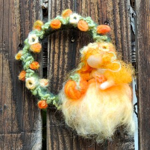 May include: A whimsical felt doll with orange and yellow hair, wearing a white dress and holding a small orange pumpkin. The doll is adorned with a wreath of green leaves and orange and white flowers.