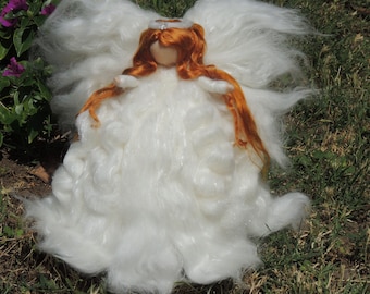 May include: A handmade angel doll with a white fluffy body and large wings. The doll has red hair and a small halo. The angel is sitting on green grass near purple flowers.
