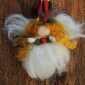 Może przedstawiać: Ręcznie robiona ozdoba świąteczna w kształcie anioła z białym, puszystym korpusem i skrzydłami. Anioł ma pomarańczowo-żółte włosy, filcową twarz i jest ozdobiony zielonym ostrokrzewem i czerwonymi jagodami. Czerwona pętla jest przymocowana do zawieszenia.