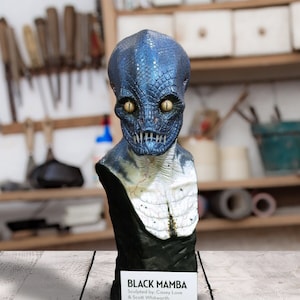 May include: A blue and black resin bust sculpture of a Black Mamba snake with yellow eyes. The sculpture is on a wooden surface with a white card that reads "BLACK MAMBA Sculpted by: Casey Love & Scott Whitworth".