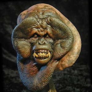 May include: A detailed, hand-painted bust of a grotesque, ape-like creature with a large, fleshy head and a menacing expression. The creature has a wide mouth with sharp teeth and bulging eyes. The bust is mounted on a metal base.