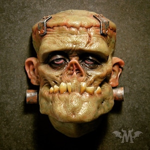 Könnte beinhalten: Detaillierte Maske von Frankensteins Monster in einem kränklichen Grünton. Die Maske weist sichtbare Nähte, Metallbefestigungen und hervorstehende Zähne auf. Die Augen sind blutunterlaufen, und das Design ist sehr detailliert.