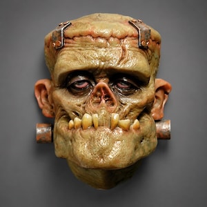 Op de afbeelding: Een sculpturaal wandmasker van een wezen met olijfkleurige, getextureerde huid, zware oogleden en een brede kaak. De figuur heeft metaalachtig uitziende bouten op het voorhoofd en de nek, samen met opvallende, onregelmatige gele tanden.