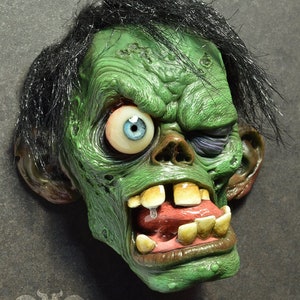 Peut inclure: Un visage vert, grotesque, semblable à celui d'un zombie, avec un œil qui sort et une bouche grande ouverte avec des dents. Le visage a une coiffure noire et hirsute.