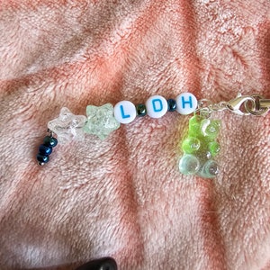 May include: A handmade keychain with a silver clasp. The keychain features clear star-shaped beads, white letter beads spelling "LDH", and a green gummy bear charm. Small iridescent beads and a silver chain complete the design. The background is a pink, fuzzy fabric.