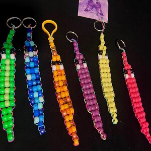 May include: Seven colorful beaded keychains shaped like worms. The keychains are made of small plastic beads in various colors, including green, blue, orange, purple, and yellow. Each keychain has a silver ring at the top.