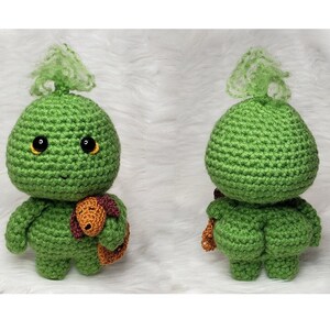 May include: A green crocheted plush toy with a brown crocheted accessory. The toy has a round head with a green tuft on top and black eyes. The toy is shown from the front and back.