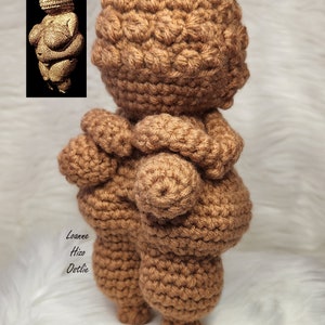 May include: A crocheted figurine of the Venus of Willendorf, a prehistoric statue of a woman, in a brown color. The figurine is standing on a white surface.