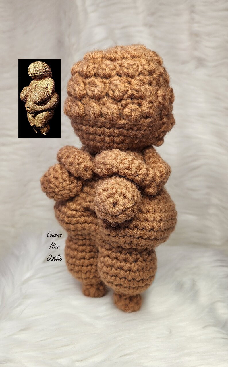 May include: A crocheted figurine of the Venus of Willendorf, a prehistoric statue of a woman, in a brown color. The figurine is standing on a white surface.