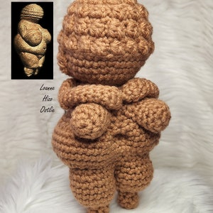 May include: Crocheted brown figurine of the Venus of Willendorf, a prehistoric statue of a woman. The figurine is standing with its arms crossed over its chest. The figurine is crocheted in a brown yarn and has a textured surface.