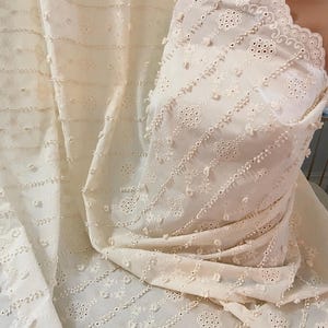 Puede incluir: Tela de algodón blanca con un delicado patrón de bordado de ojales. La tela tiene una superficie texturizada y un borde festoneado.