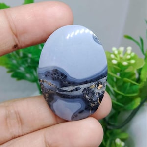 Op de afbeelding: Ovale edelsteen met een lichtblauw bovenste gedeelte en een onderste gedeelte met een donker, gelaagd patroon met gouden vlekken. De gepolijste steen wordt in een hand gehouden, wat de natuurlijke schoonheid benadrukt.