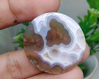 May include: A circular gemstone with a distinctive pattern. The stone displays concentric bands of white, brown, and tan, creating a marbled effect. The gemstone is held between fingers, highlighting its natural beauty.